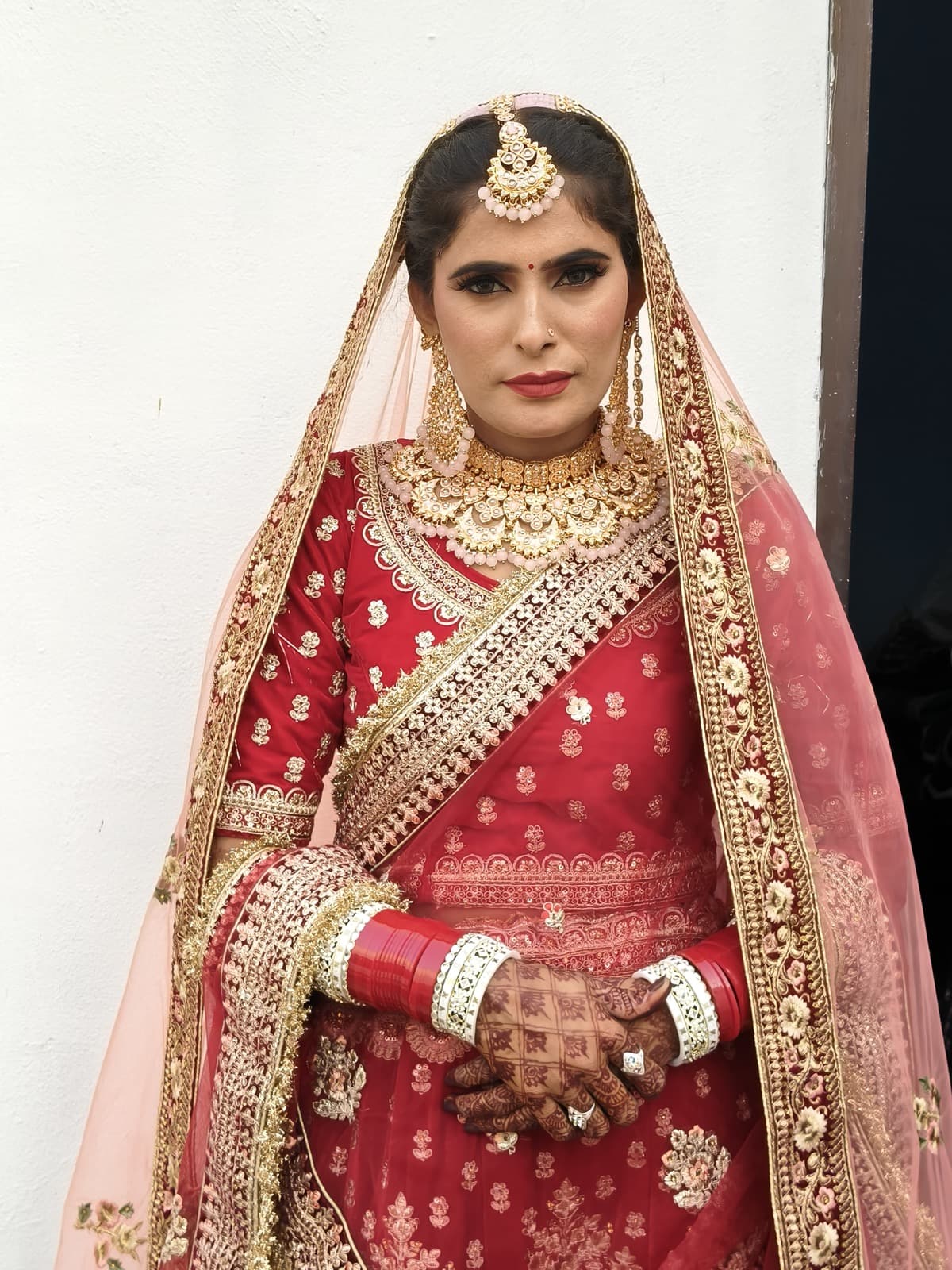 Classic bridal makeup with red lips, heavy gold kundan jewellery, and veil on a bride in a red and gold embroidered lehenga