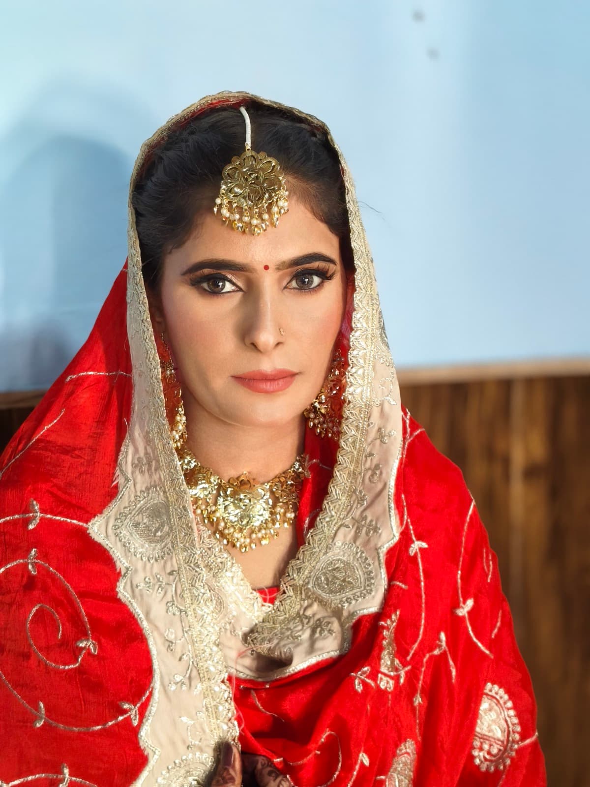 Traditional bridal makeup with intense eyes, bold brows, gold maang tikka, and red dupatta on a bride in a red banarasi outfit