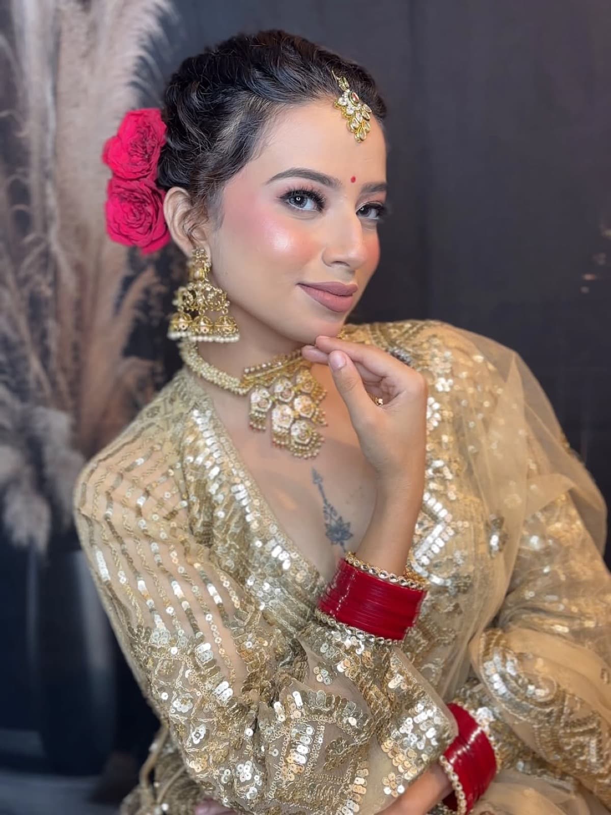Bridal close-up with glowing dewy skin, soft nude eyes, pink rose hair accessory, and layered gold kundan jewellery