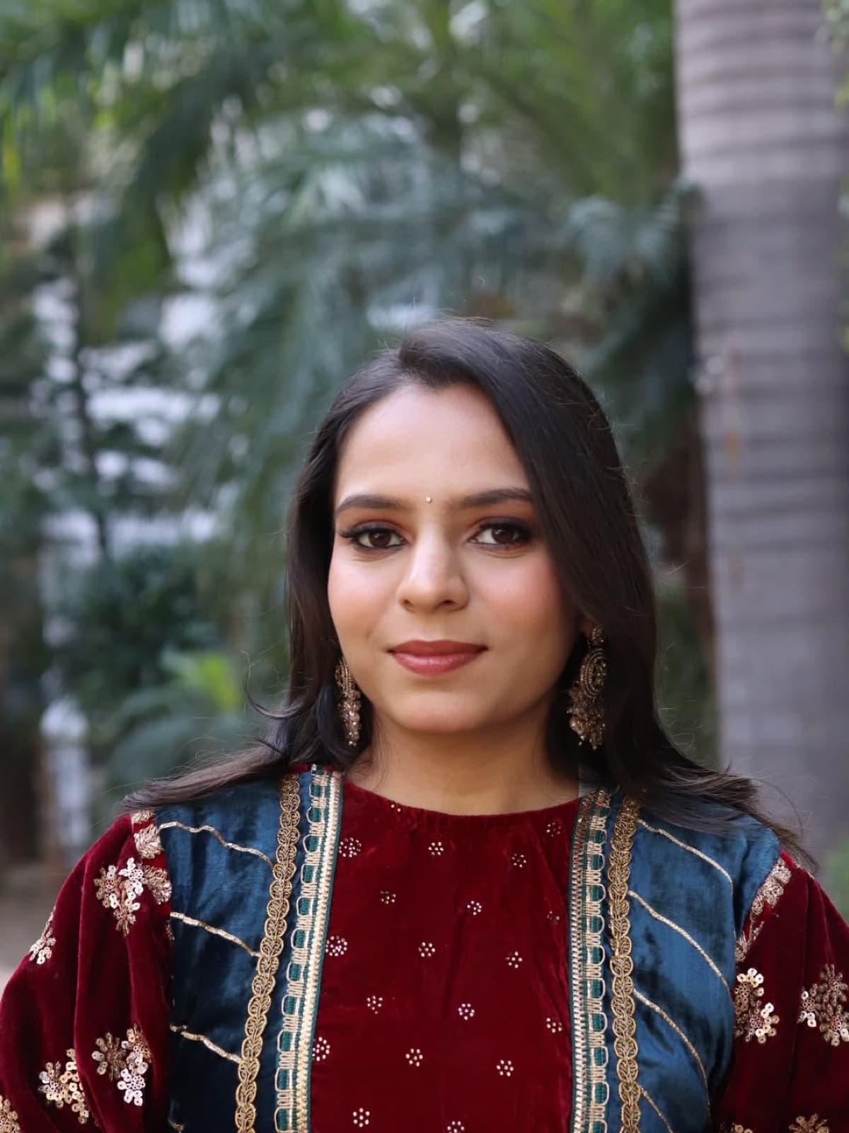 Minimal natural makeup look with clean skin, bindi, and gold jhumka earrings on a client in a maroon velvet suit, garden setting