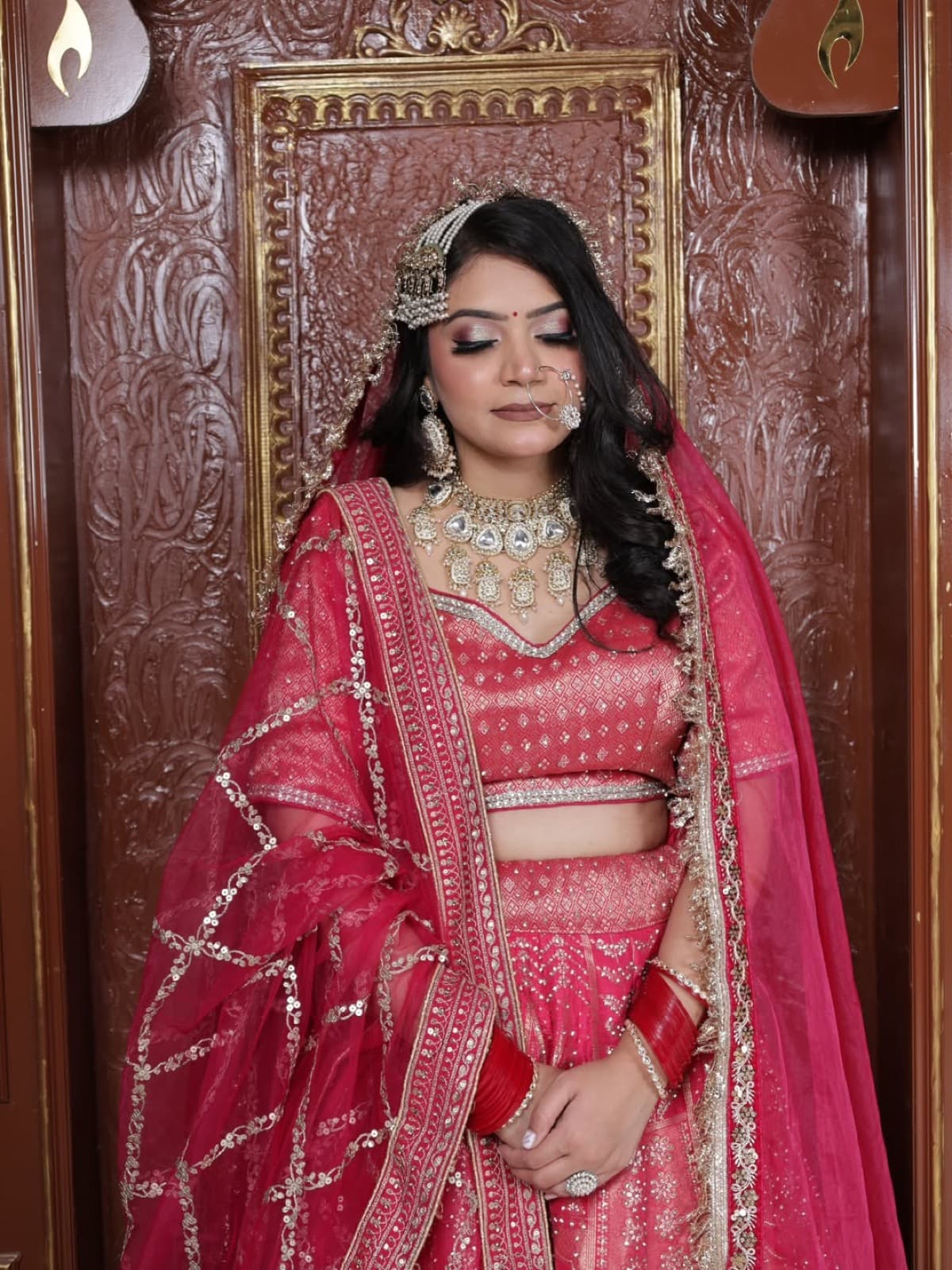 Full bridal portrait with soft glam makeup on a bride in a pink embroidered lehenga with gold necklace, eyes downcast, ornate wooden backdrop