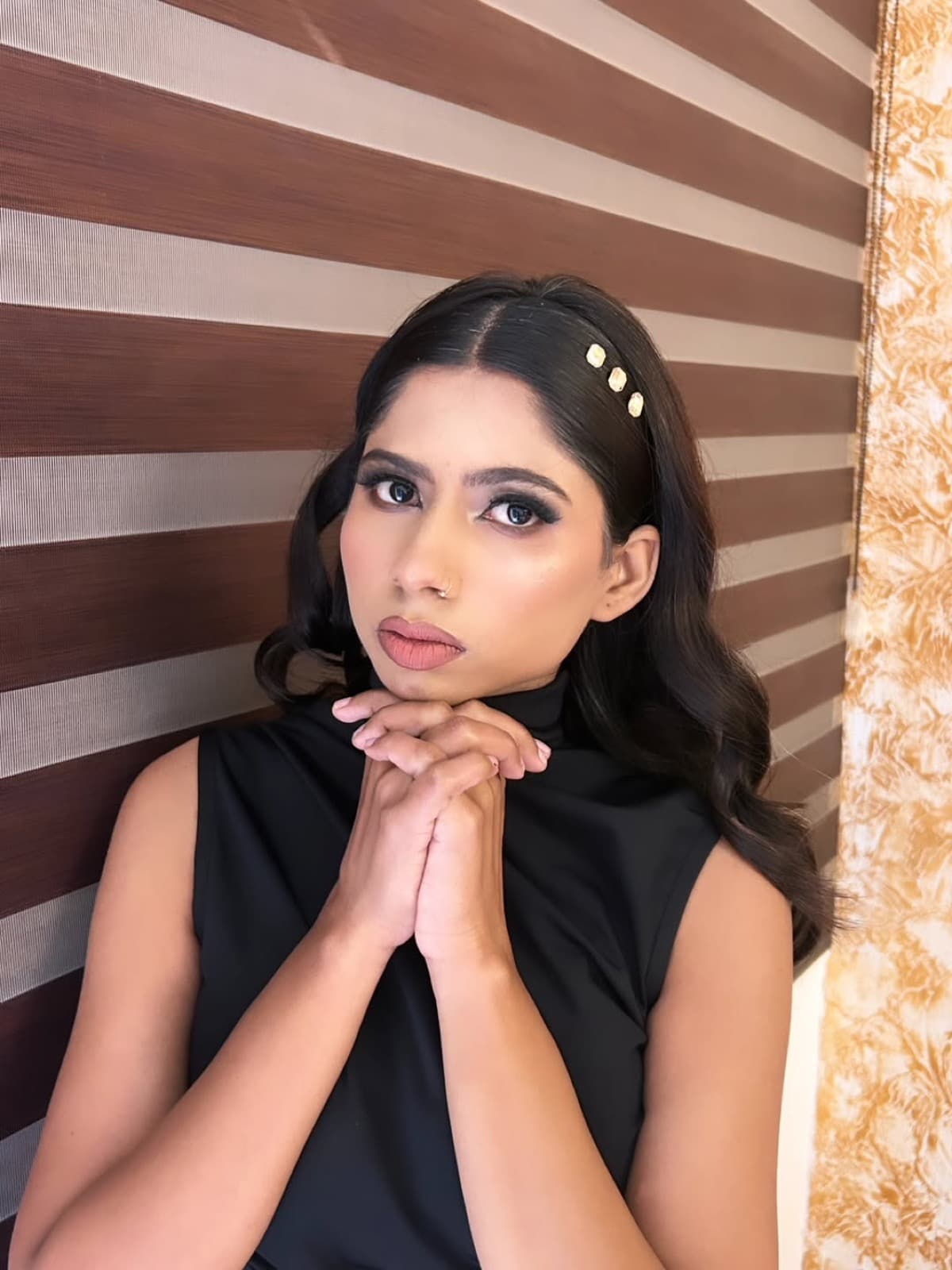 Dramatic smokey eye with matte nude lips, gold hair accessories, and soft curls on a model in a black sleeveless top, indoor portrait