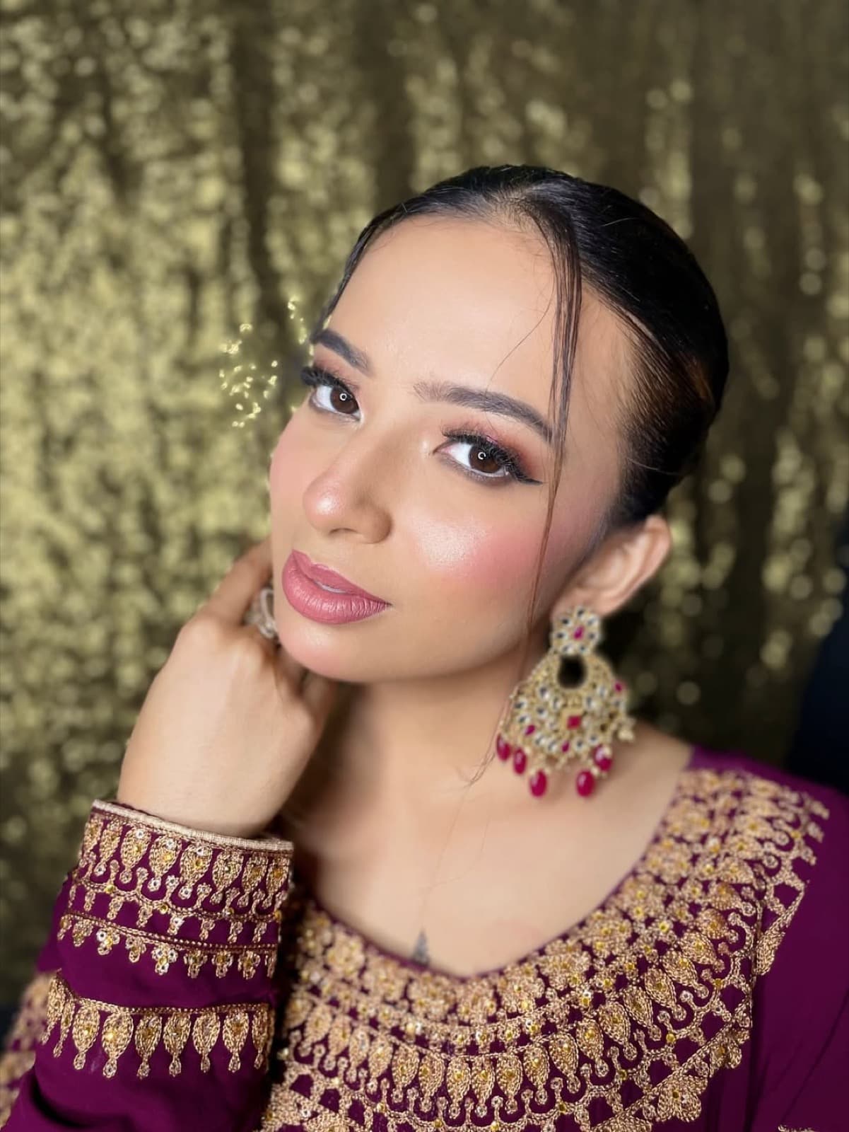 Close-up glam with highlighted cheekbones, soft pink lips, kundan earrings with ruby drops, and sleek bun on a model in plum and gold outfit