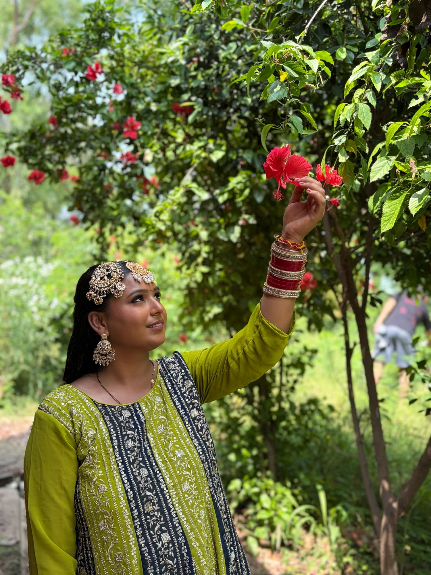 Outdoor mehndi portrait in a lime green outfit with gold jewellery, reaching for a red flower