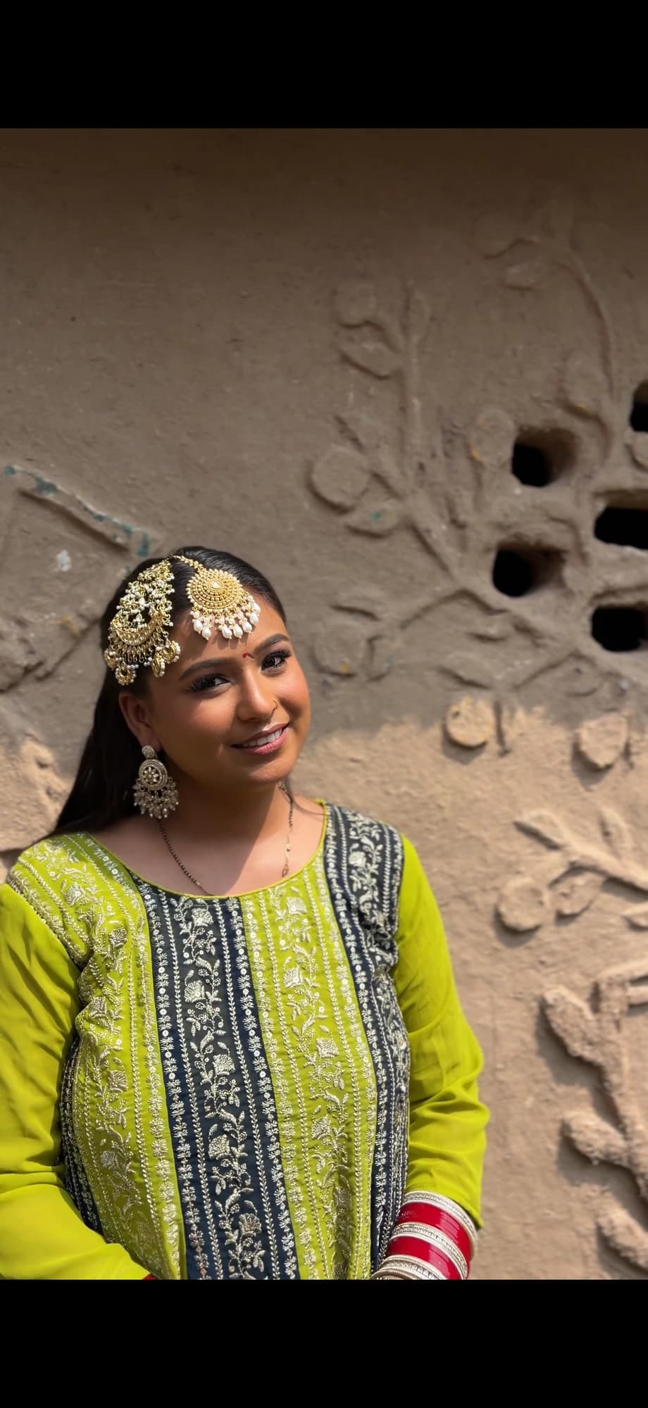 Mehndi makeup with softly defined eyes and pink lips, wearing a lime green embroidered outfit beside a carved wall
