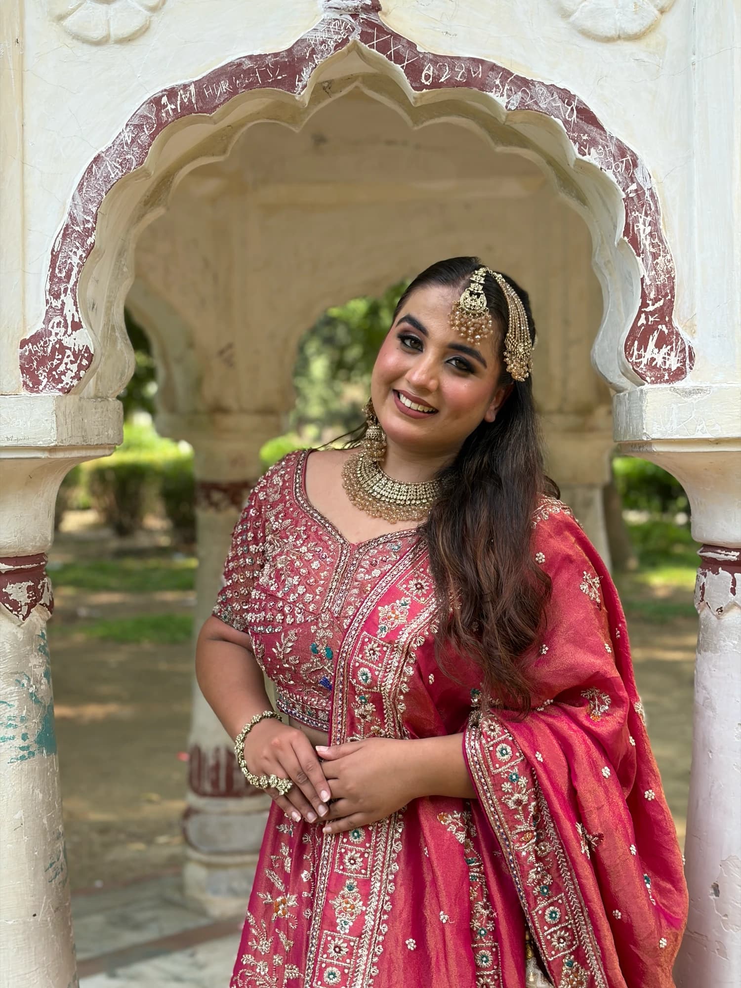 Smiling portrait with soft eye makeup and a pink embroidered outfit, framed by a decorative garden arch