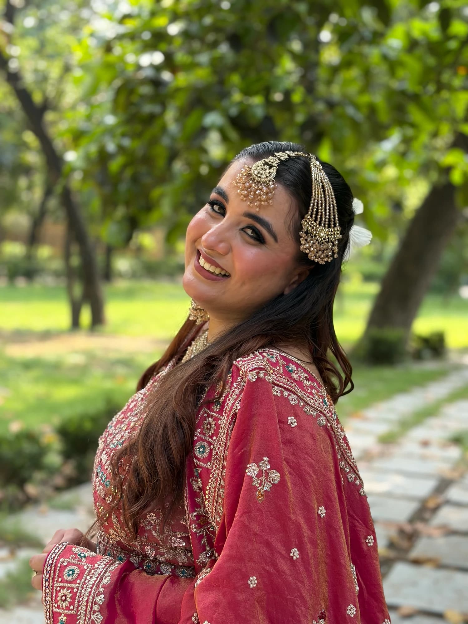 Over-the-shoulder garden portrait with softly glowing makeup, loose waves and a pink embroidered outfit