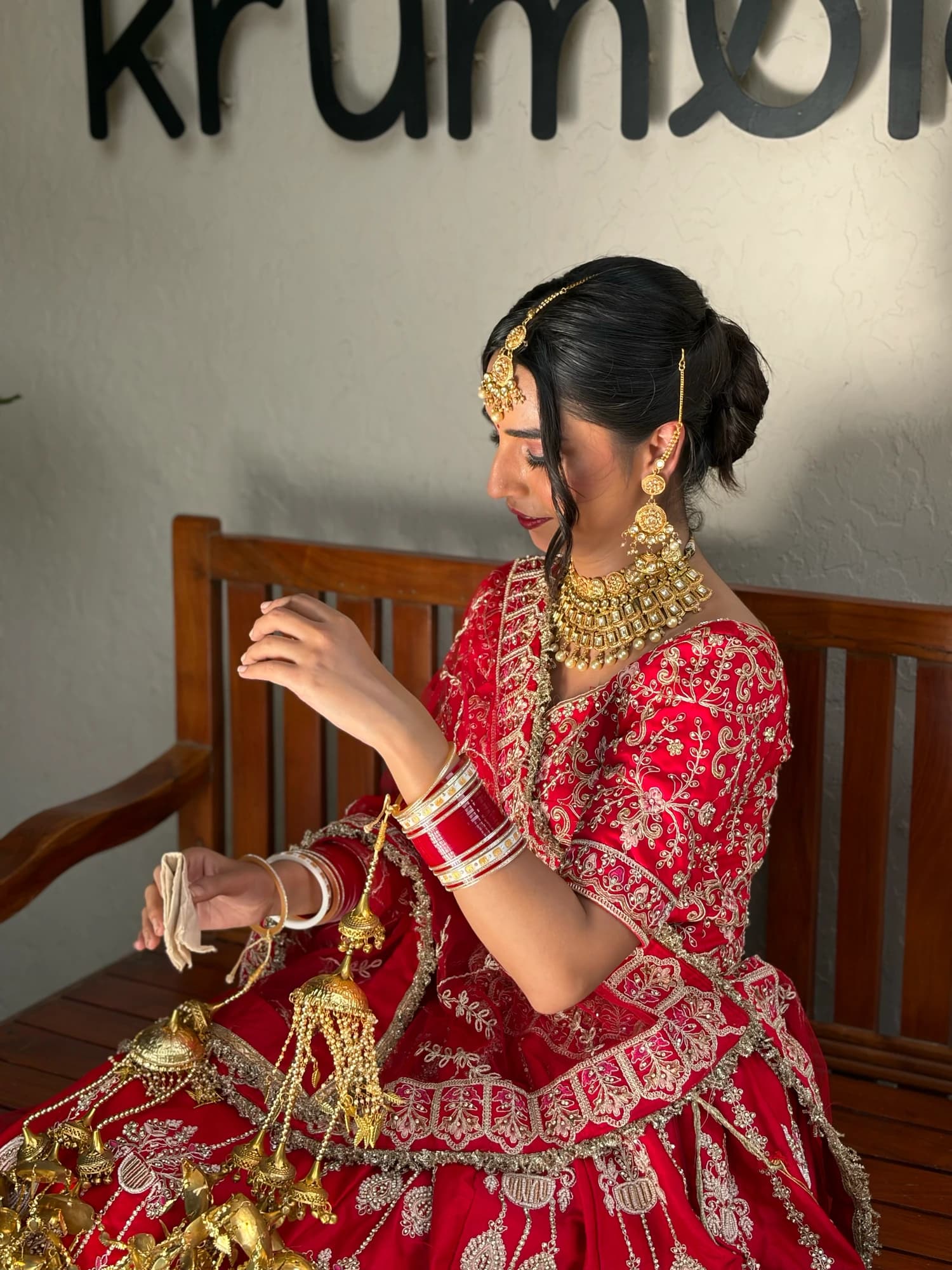 Seated bridal side portrait with defined eyes, a sleek bun, gold jewellery and a red embroidered lehenga