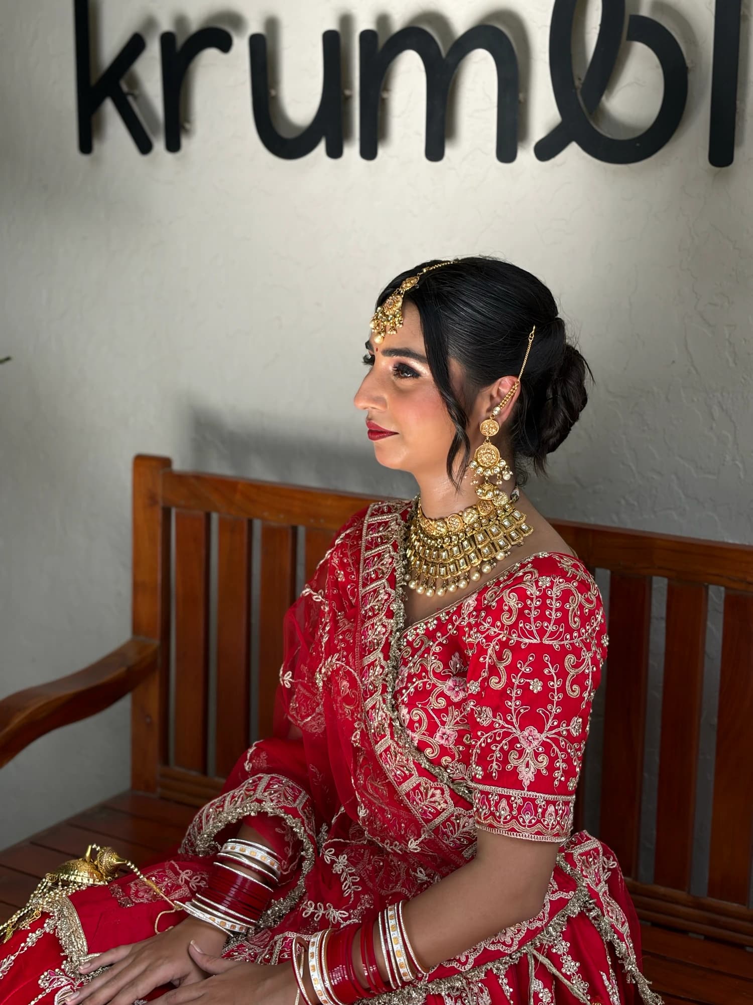 Bridal profile with warm eye makeup, rose lips and a sleek bun, seated in a red and gold lehenga