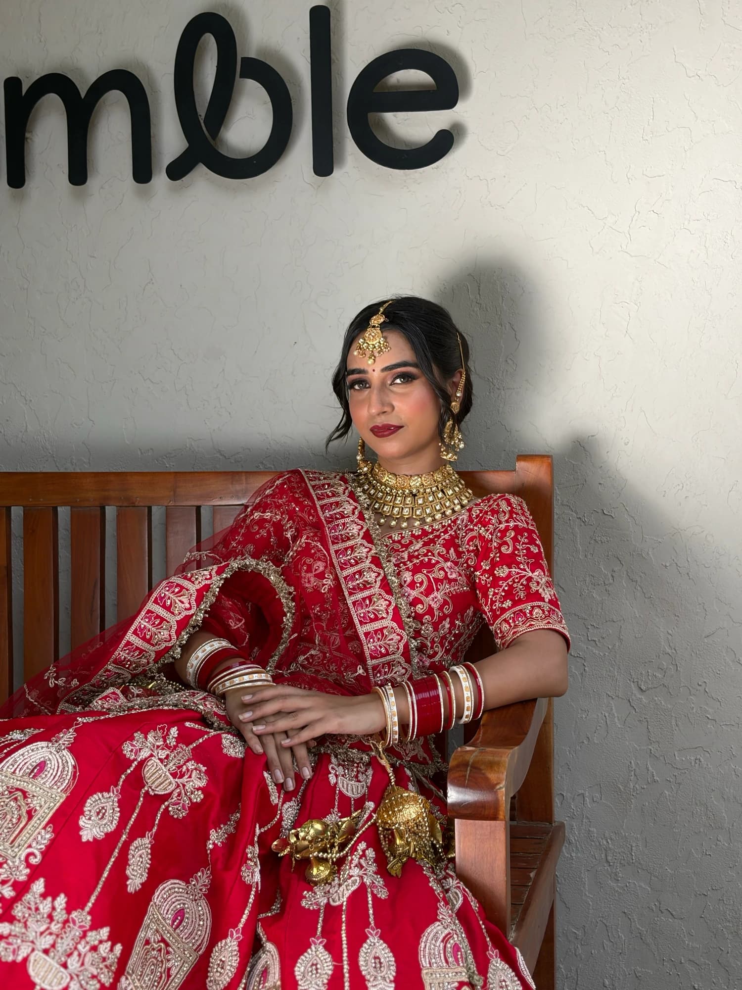 Seated bridal portrait with glowing skin, defined eyes and layered gold jewellery, wearing a red lehenga