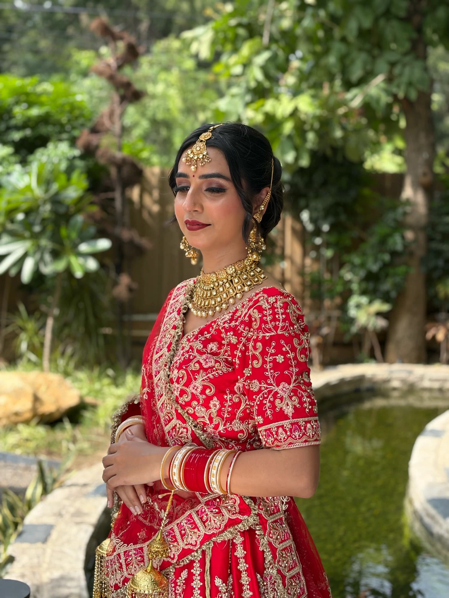 Outdoor bridal profile with softly defined eyes, rose lips and gold jewellery against a red embroidered lehenga