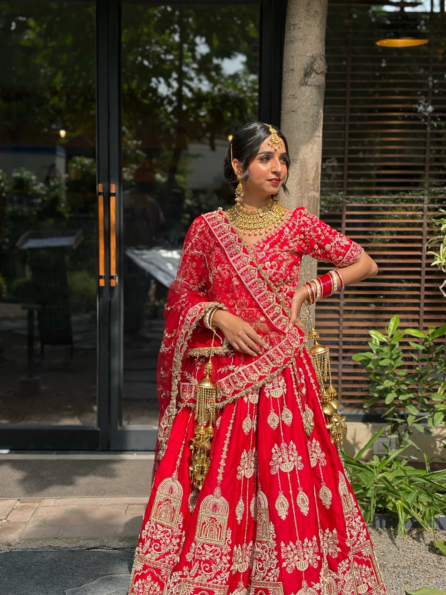 Full-length bridal portrait in a red embroidered lehenga with layered gold jewellery, standing beside a glass doorway