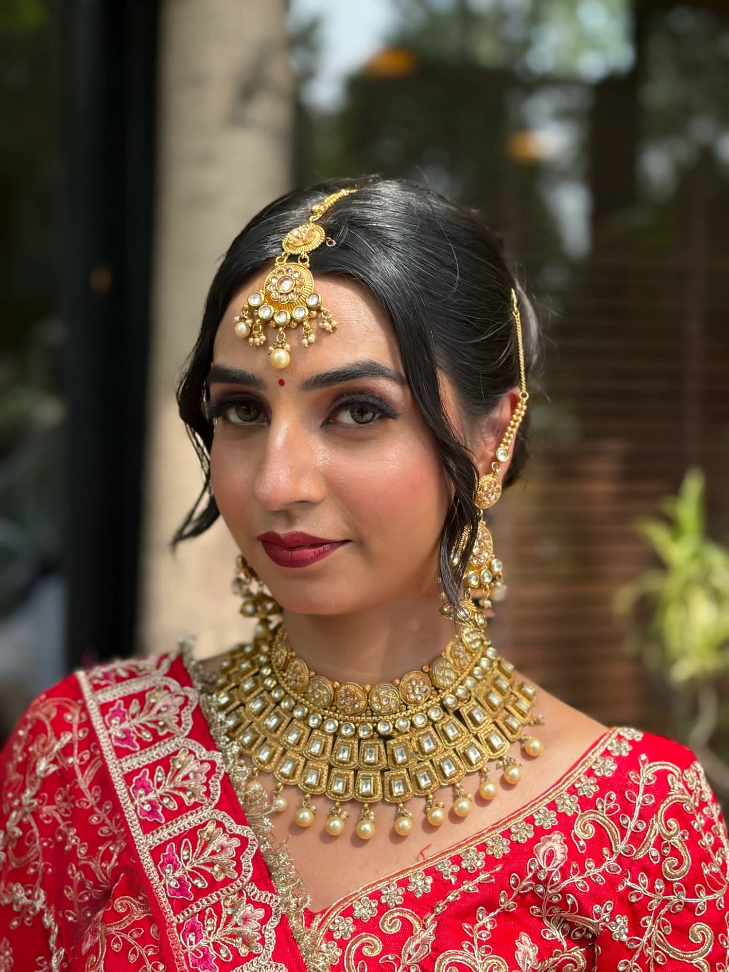 Front-facing bridal close-up with defined eyes, rose lips, a gold maang tikka and layered gold jewellery