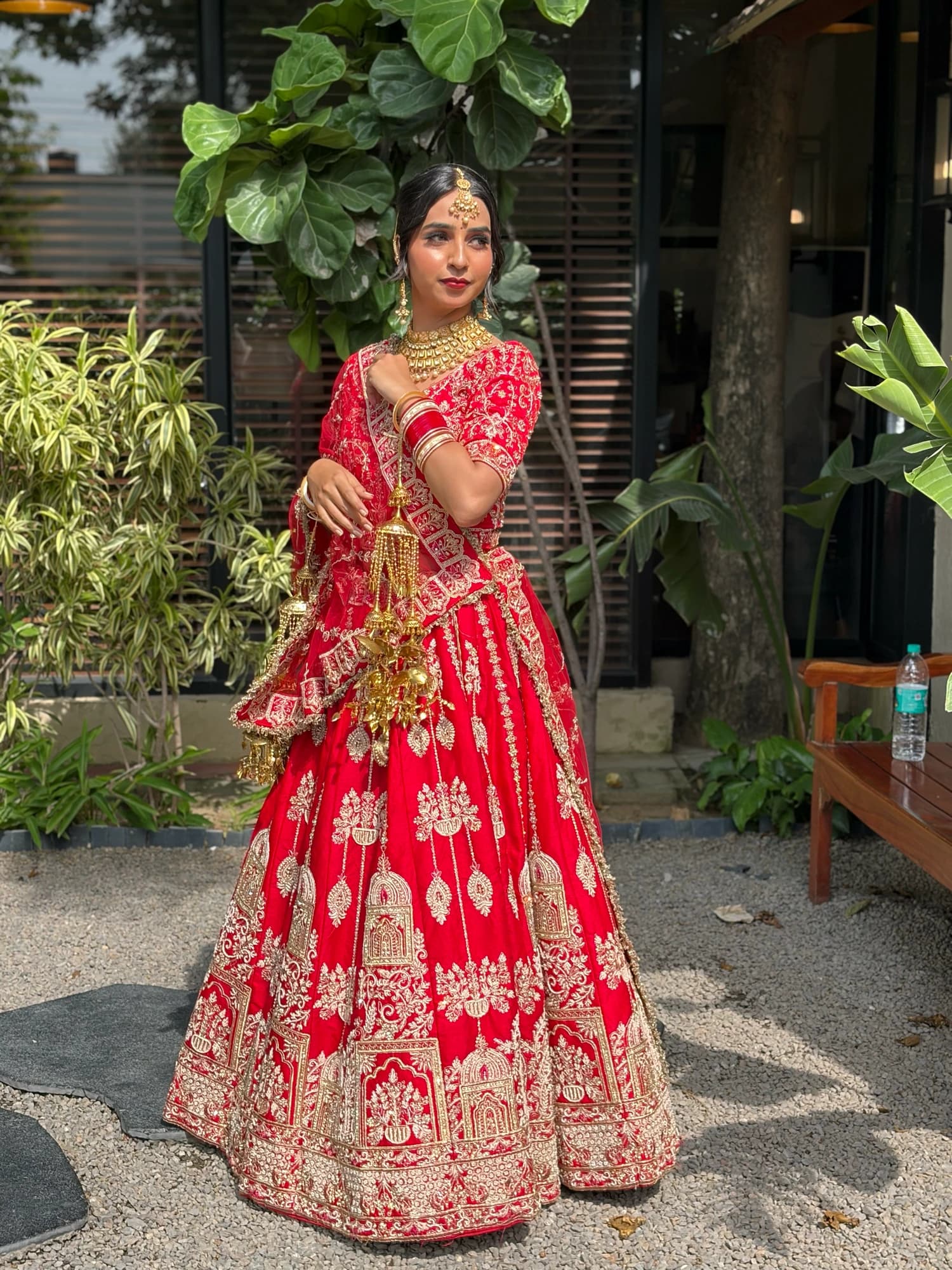 Full-length bridal look with a red embroidered lehenga, gold kaleere and a sleek bun beside garden greenery