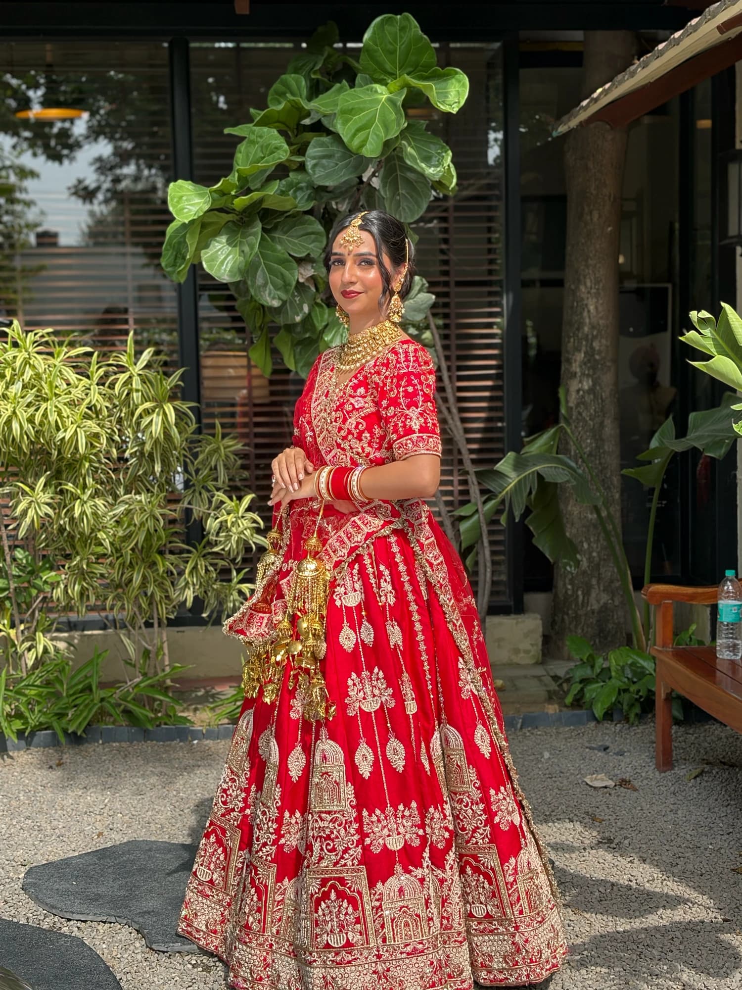 Full-length outdoor bridal portrait with glowing makeup, a red and gold lehenga and traditional gold jewellery