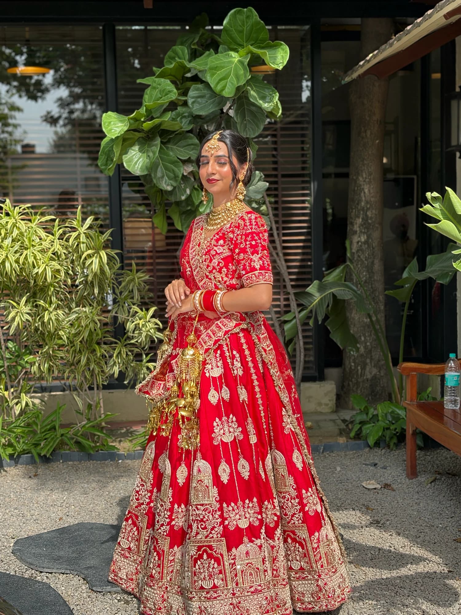 Outdoor bridal pose with softly defined makeup, gold jewellery and a red embroidered lehenga beside a glass doorway