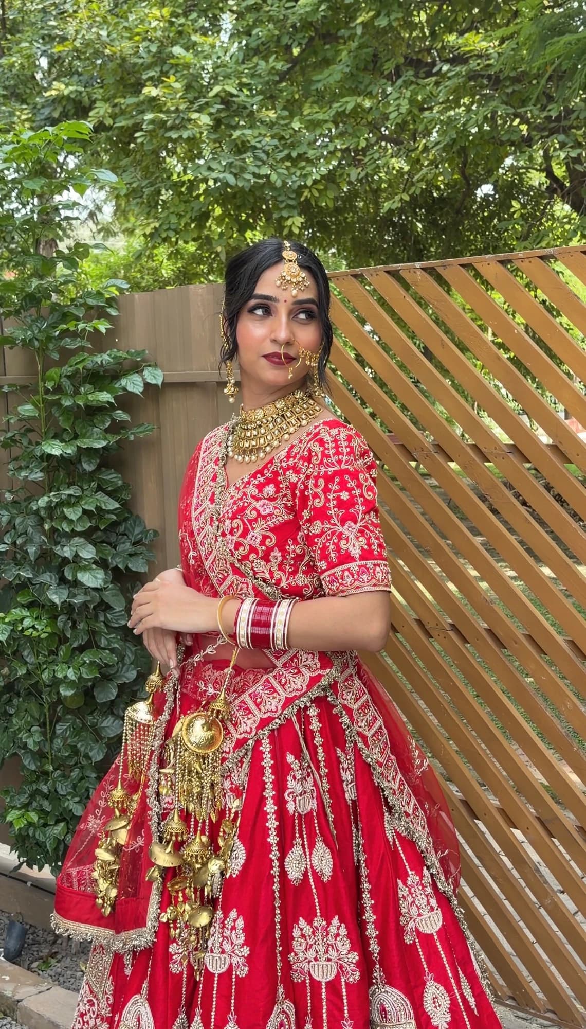 Bridal portrait with defined eyes, rose lips, a traditional nose ring and red lehenga against garden greenery