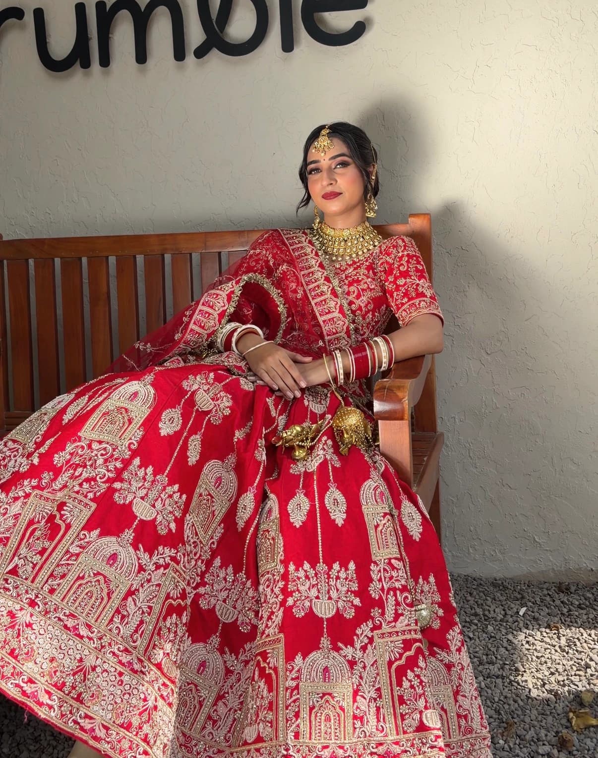 Seated bridal portrait with a red embroidered skirt spread across a wooden bench and softly defined makeup