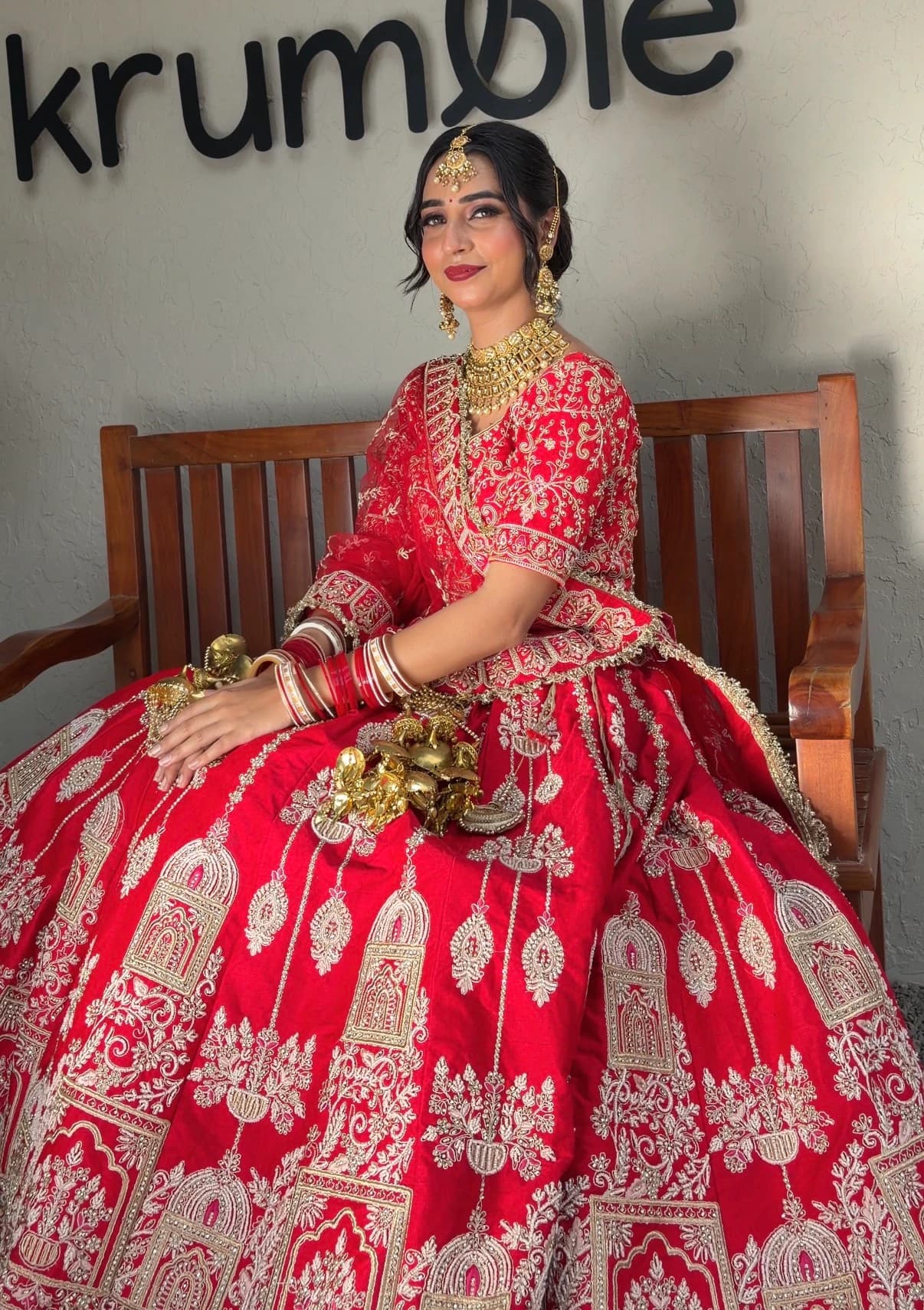 Seated bridal side portrait with glowing skin, gold jewellery and a red lehenga on a wooden bench